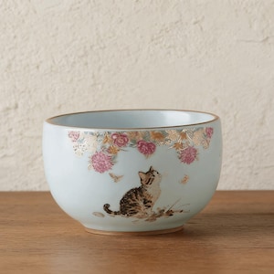 May include: A light blue ceramic bowl with a gold rim and a decorative floral and cat design. The bowl features a brown and white tabby cat looking upwards, with pink flowers and butterflies. The bowl is a decorative item.