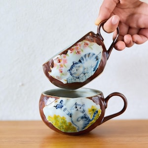 Puede incluir: Dos tazas de cerámica con bordes y asas marrones. Cada taza presenta un diseño pintado a mano de un gato. Un gato está durmiendo entre flores rosas, y el otro está alcanzando una mariposa azul. Las tazas están sobre una superficie de madera.