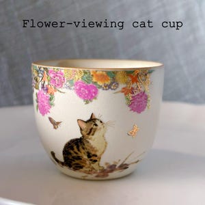 May include: A white ceramic cup with a floral design and a cat illustration. The cup features a gold rim and the text "Flower-viewing cat cup". The cat is looking up at a butterfly.