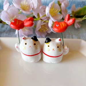 May include: Two white ceramic cat figurines with black hats and red accents, holding red and orange fish ornaments. The figurines have a red stripe around the base and are set against a backdrop of white flowers.