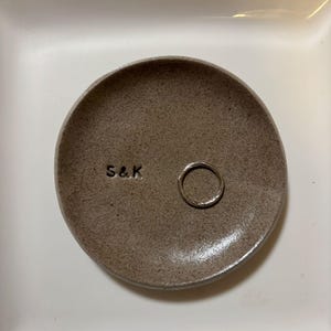 Peut inclure: Un petit plat à bijoux rond en céramique grise mouchetée avec les initiales "S&K" estampillées sur la surface. Une bague en argent repose à l'intérieur du plat. Le plat est posé sur une surface blanche.