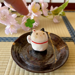May include: A ceramic Maneki Neko cat figurine, white with black and yellow accents, holding a green four-leaf clover. The cat sits on a dark brown plate with gold accents. Decorative cherry blossoms are in the background.