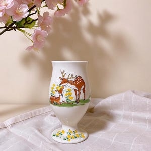May include: A white ceramic pedestal vase featuring a colorful illustration of two brown spotted deer standing and sitting among yellow flowers and green grass. The base of the vase is also decorated with a pattern of small yellow flowers and green leaves.