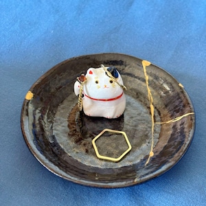 May include: A small, round, dark brown ceramic dish with gold accents and a white lucky cat figurine. A gold hexagonal ring rests on the dish. The cat has a red collar and a gold chain. The dish has a Kintsugi repair design.