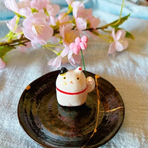May include: A white and black ceramic Maneki Neko cat figurine with a red collar, holding a pink flower. The cat sits on a black plate with gold accents, with pink cherry blossoms in the background.