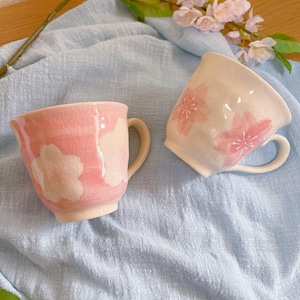 May include: Two ceramic mugs with floral designs. One is pink with white floral accents, the other white with pink floral accents. Both have a curved handle and sit on a light blue cloth.