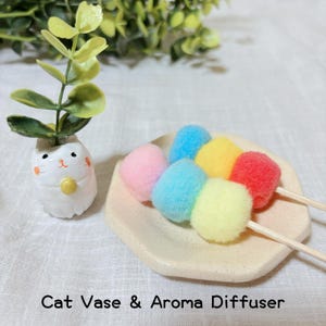 Puede incluir: Un jarrón de cerámica blanca con forma de gato que sostiene una ramita de hojas verdes, junto a una bandeja hexagonal beige con dos brochetas de pompones coloridos y difusos en rosa, azul, amarillo y rojo. Se ve el texto Cat Vase & Aroma Diffuser.