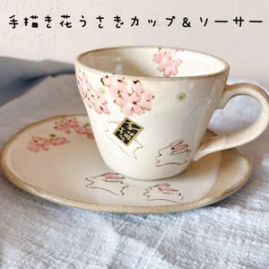May include: A ceramic teacup and saucer set. The cup and saucer are off-white with hand-painted pink cherry blossoms and green dots. The cup has a handle and a small square emblem. The saucer features bunny illustrations.