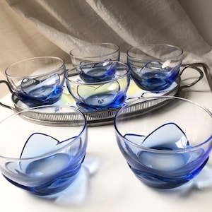 Pode incluir: Um conjunto de seis chávenas e pires de vidro transparente com um design de ondas azuis. As chávenas têm pegas prateadas e assentam numa bandeja prateada. As chávenas e pires estão dispostos numa superfície branca.