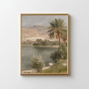 May include: A framed landscape painting with a gold frame. The artwork depicts a serene scene with a body of water, palm trees, and distant mountains under a cloudy sky. The color palette is muted, with earthy tones and soft greens.