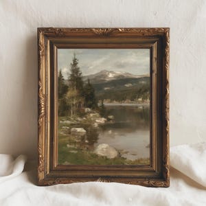 以下が含まれることがあります： 湖の風景と背景に山々が描かれた額入りの風景画。このアートワークは、金色の装飾フレームが特徴です。絵画には、木々、岩、穏やかな水面、そして曇り空が描かれています。