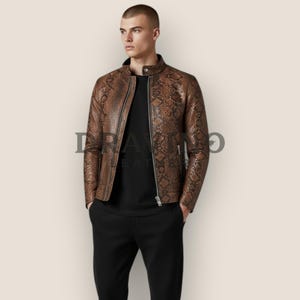 May include: A brown snakeskin-patterned leather jacket with a stand-up collar, a full-length silver zip, and long sleeves. The jacket features the text "DRAVINO LEATHER" printed across the chest area. It is worn over a plain black shirt.