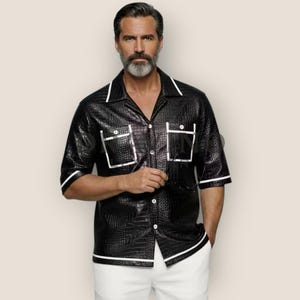 Può includere: Una camicia a maniche corte abbottonata di colore nero con un motivo testurizzato in pelle di coccodrillo. La camicia presenta un colletto a revers, due tasche sul petto con bordi bianchi e profili bianchi lungo il colletto, le tasche e i polsini.