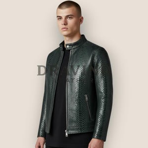 Può includere: Una giacca verde scuro con motivo testurizzato in pelle di serpente, colletto alla coreana con chiusura a bottone automatico e cerniera frontale color argento. La giacca presenta una tasca con zip sul lato sinistro sopra una maglia nera.