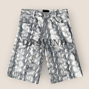 May include: High-waisted shorts featuring a grey and white snakeskin pattern. The garment includes a front button closure, belt loops, and two front pockets. The text "DRAVINO LEATHER" is printed in dark grey across the centre of the shorts.