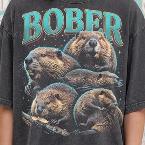 May include: Dark gray t-shirt featuring a graphic of several beavers with the word "BOBER" in teal letters. The text "PEPPER COMFORT COLORS" is printed at the bottom of the shirt.