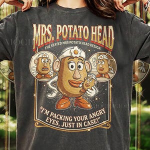 May include: Dark gray t-shirt featuring a Mrs. Potato Head design. The design includes the text "MRS. POTATO HEAD" and "I'M PACKING YOUR ANGRY EYES, JUST IN CASE!" in a vintage style. The shirt is labeled "PEPPER COMFORT COLORS".