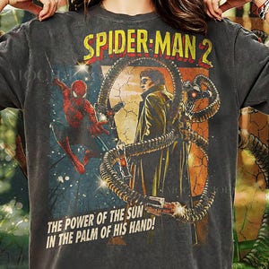 Puede incluir: Camiseta gris oscuro con un gráfico de Spider-Man 2. El diseño incluye a Spider-Man, Doctor Octopus y el texto "SPIDER-MAN 2" y "THE POWER OF THE SUN IN THE PALM OF HIS HAND!". La camiseta está etiquetada como "PEPPER COMFORT COLORS".