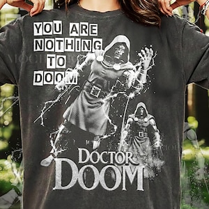 May include: Dark gray t-shirt featuring a graphic of Doctor Doom with the text "YOU ARE NOTHING TO DOOM" and "DOCTOR DOOM". The shirt is printed with white ink and has a vintage, distressed look. The words "PEPPER COMFORT COLORS" are at the bottom.