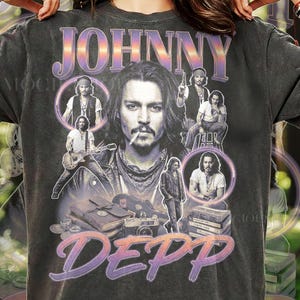Könnte beinhalten: Ein anthrazitfarbenes Kurzarm-T-Shirt mit einem großen Grafikdruck von Johnny Depp. Das Design zeigt mehrere Schwarz-Weiß-Fotos des Schauspielers, eine Gitarre, eine Kamera und Bücher sowie den Schriftzug JOHNNY DEPP in einer Farbverlaufsschrift.