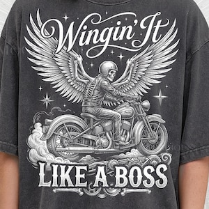 May include: Dark gray t-shirt featuring a detailed graphic of a skeleton biker riding a motorcycle with angel wings. The text "Wingin' It" and "Like a Boss" are displayed in white. The shirt is from Pepper Comfort Colors.
