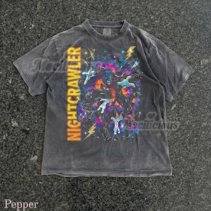 Op de afbeelding: Donkergrijs t-shirt met de gele tekst "NIGHTCRAWLER". De afbeelding toont een blauwhuidig personage met uitgestrekte handen tegen een levendige, wervelende achtergrond en bliksem.