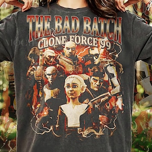 Pode incluir: T-shirt cinzenta escura com o texto "THE BAD BATCH CLONE FORCE 99" em vermelho e dourado. A camisa apresenta um gráfico de personagens da série animada, com detalhes de raios. Feita de Pepper Comfort Colors.