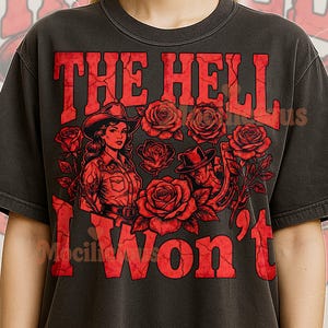 Puede incluir: Camiseta gris oscuro con un gráfico rojo desgastado. El gráfico presenta el texto "THE HELL I WON'T" sobre un diseño de rosas y dos figuras con sombreros de vaquero. La camiseta es de estilo vintage.
