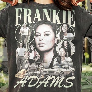 May include: A charcoal grey t-shirt featuring a vintage-style graphic print of Frankie Adams. The design includes multiple portraits of the individual, a classic car, film reels, and cameras, with the text FRANKIE ADAMS displayed in large lettering.