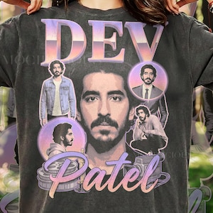 May include: A charcoal gray t-shirt featuring a large graphic print of actor Dev Patel. The design includes a central portrait, three smaller circular images of the actor, a film reel, a trophy, and the text DEV PATEL in a gradient purple font.