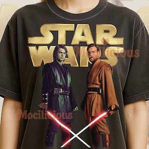 May include: Dark gray t-shirt featuring the Star Wars logo in gold, with images of Anakin Skywalker and Obi-Wan Kenobi holding lightsabers. The shirt has a vintage look and the text "Mocilicious" is printed on the shirt.