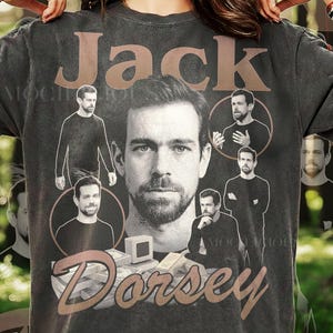 Peut inclure: Un t-shirt gris anthracite avec une grande impression graphique de Jack Dorsey. Le motif comprend plusieurs portraits photographiques en noir et blanc de l'individu, le nom Jack Dorsey en grandes lettres marron et une pile de billets en bas.