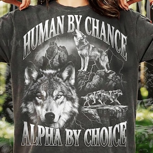 May include: Dark gray t-shirt featuring a detailed wolf graphic. The design includes a close-up wolf face, a howling wolf, and running wolves, with the text "HUMAN BY CHANCE" and "ALPHA BY CHOICE" in white.