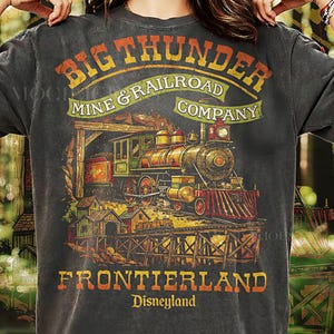 May include: Dark grey t-shirt with a vintage train illustration. Text reads "BIG THUNDER MINE & RAILROAD COMPANY" and "FRONTIERLAND Disneyland". The design features a train, wooden bridge, and buildings, in a retro style.