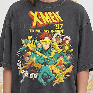 Può includere: T-shirt grigio scuro con una grafica colorata dei personaggi degli X-Men. Il design include il testo "X-Men '97" e "To Me, My X-Men". La maglietta mostra vari personaggi dei fumetti in pose dinamiche.