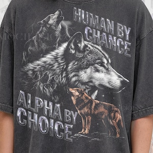 May include: Dark gray t-shirt featuring a detailed graphic of three wolves, with the text "HUMAN BY CHANCE" and "ALPHA BY CHOICE" in a reflective, silver font. The shirt is from Pepper Comfort Colors.
