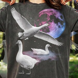 Pode incluir: Camiseta cinza escuro com um design celestial. Apresenta gansos brancos, uma lua vibrante e estrelas. Um ganso está em voo, e dois estão no chão ou na água. O design tem um estilo artístico e sonhador.