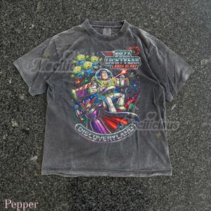 Puede incluir: Una camiseta gris oscuro con un gráfico colorido de Buzz Lightyear y otros personajes de Toy Story. El gráfico incluye el texto "Buzz Lightyear Laser Blast" y "Discoveryland".