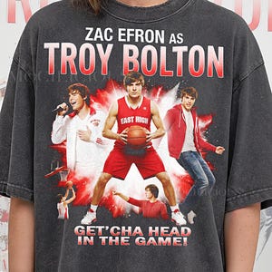 May include: Dark gray t-shirt featuring Zac Efron as Troy Bolton from High School Musical. The design includes multiple images of the character, the text "ZAC EFRON AS TROY BOLTON", and the phrase "GET'CHA HEAD IN THE GAME!" in red and white.