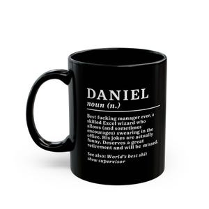 May include: Black ceramic mug with the name "DANIEL" in white, along with a humorous definition. The definition describes a manager who allows swearing and is a skilled Excel wizard. The mug has a curved handle.