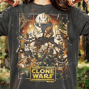 May include: Dark gray t-shirt featuring a graphic from "Star Wars: The Clone Wars." The design includes various clone troopers, vehicles, and the show's logo in gold. The text "Pepper Comfort Colors" is printed at the bottom.