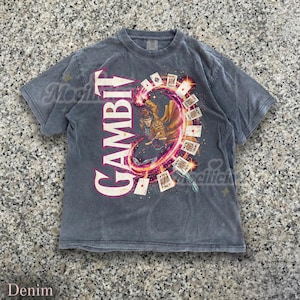 Puede incluir: Camiseta de color denim con un diseño del personaje Gambit de Marvel Comics. El diseño incluye la palabra "GAMBIT" en letras grandes y verticales, y una disposición circular de naipes. La camiseta tiene un aspecto vintage y desgastado.