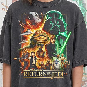 Puede incluir: Camiseta gris oscuro con un gráfico de Star Wars "Return of the Jedi". El diseño incluye a Darth Vader, Luke Skywalker con un sable de luz, Jabba the Hutt y otros personajes de la película. El texto "Pepper Comfort Colors" está en la parte inferior.