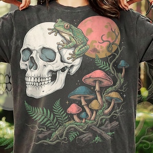 May include: Dark gray t-shirt featuring a skull with a frog on top, a pink moon, and colorful mushrooms. The design includes fern leaves and a whimsical, nature-inspired illustration.