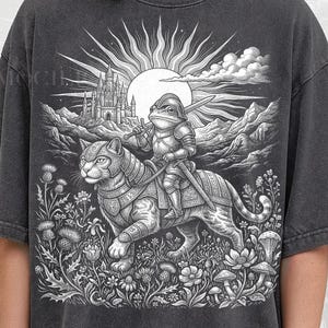 May include: Dark gray t-shirt featuring a detailed black and white illustration. A frog knight in armor rides a cat through a landscape with a castle, sun, and clouds. The text "PEPPER COMFORT COLORS" is at the bottom.