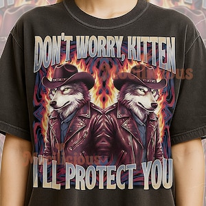 May include: Dark gray t-shirt with a graphic featuring two wolf-like figures wearing cowboy hats and leather jackets, set against a fiery background. Text reads "DON'T WORRY, KITTEN I'LL PROTECT YOU."