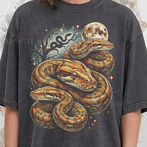 May include: Dark gray t-shirt featuring a detailed illustration of three snakes in shades of brown and gold, set against a night sky with a full moon and stars. The text "PEPPER COMFORT COLORS" is printed at the bottom.