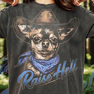 May include: Dark gray t-shirt featuring a chihuahua wearing a cowboy hat and a blue paisley bandana. The graphic includes the text "Raise Hell" in a blue, vintage-style font. The dog's portrait is detailed and realistic.