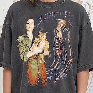 May include: Dark grey t-shirt with a graphic of a person holding a cat, alongside a futuristic machine design. The person wears a green and orange jumpsuit. The text "PEPPER COMFORT COLORS" is at the bottom of the shirt.