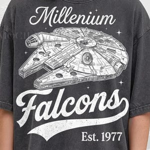 May include: Dark gray t-shirt featuring a detailed white illustration of the Millennium Falcon spaceship. The text "Millennium Falcons" and "Est. 1977" are printed in white. The shirt is from Pepper Comfort Colors.